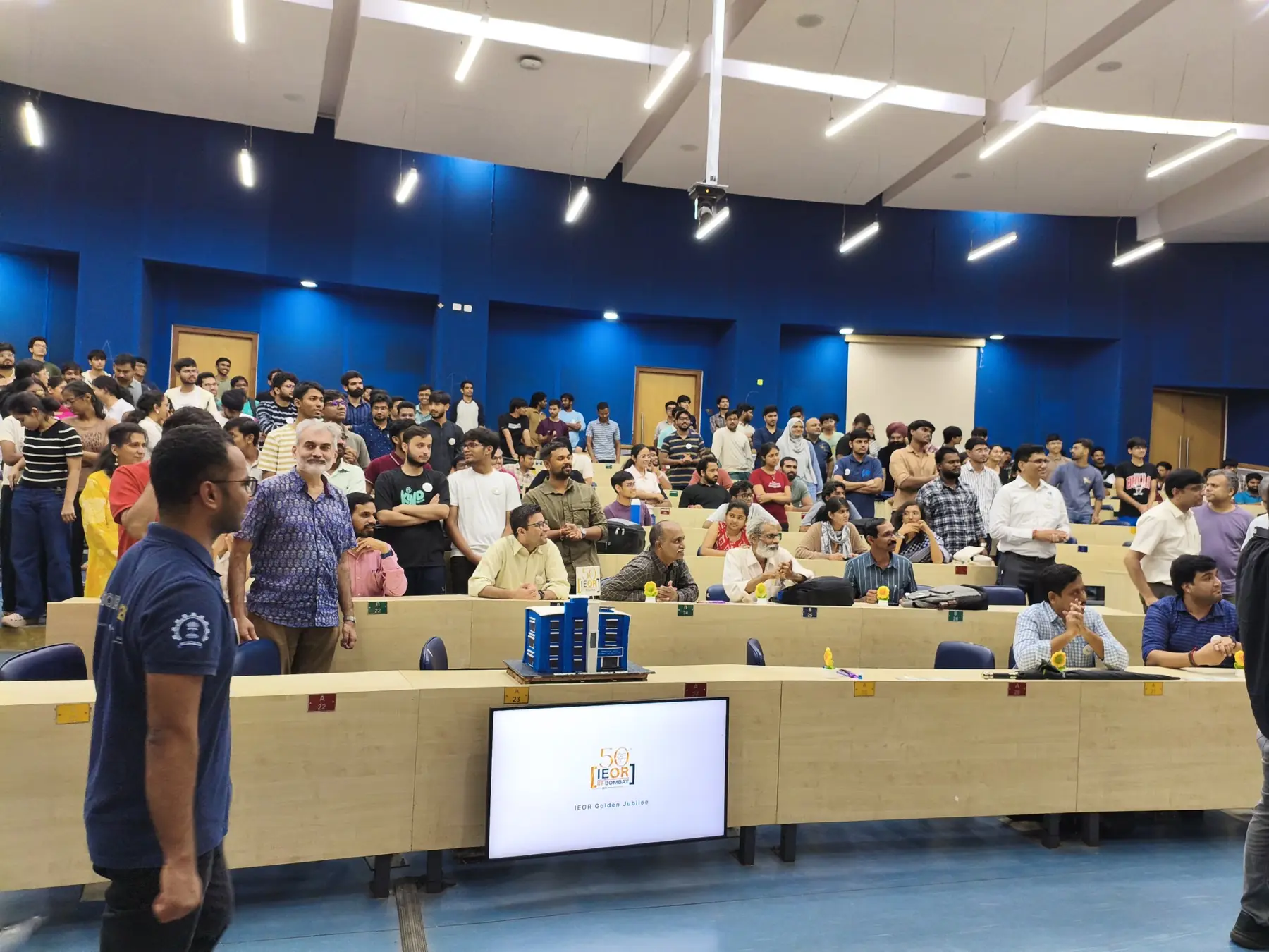 Students standing along the back of a blue lecture hall, with faculty seated in front