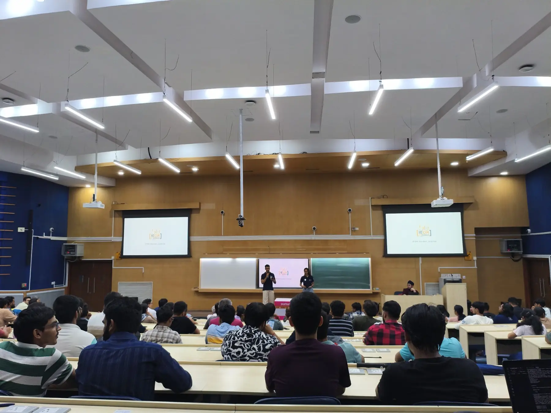 A full lecture hall seen from the back, with the Golden Jubilee mark on two screens