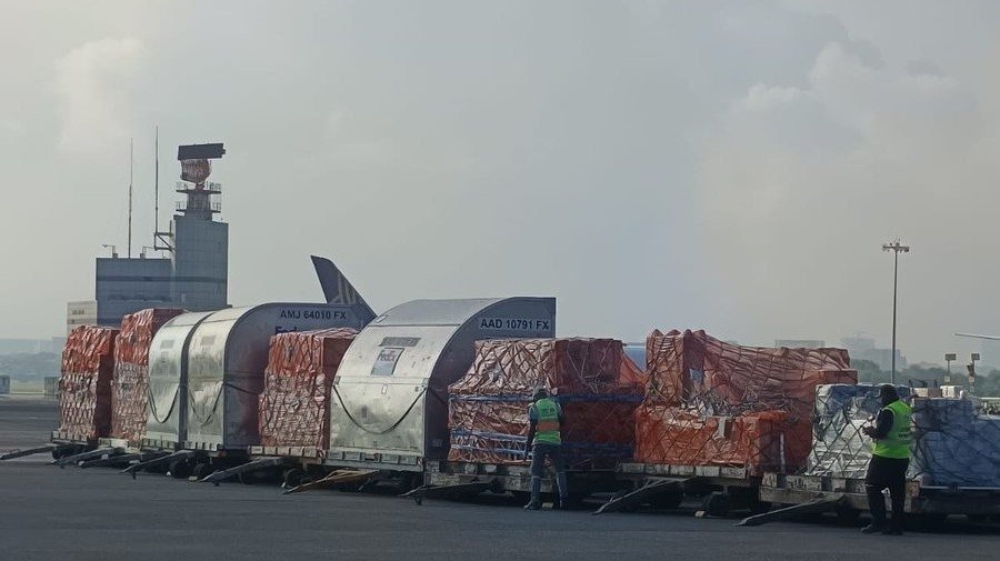 Air cargo containers lined up on an airport apron, with ground crew and the control tower behind